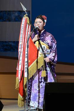 紫色の着物を着た女性が優勝旗を持って歌っている写真