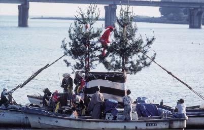 船の中に祭りで使う木の置物が置かれていて、赤い服を着た男性が木に登っている写真