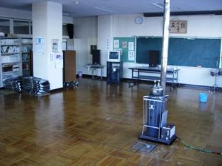 黒板、テレビ、ストーブ、バケツ、折りたたんで積み重ねられたパイプ椅子が置いてある教室内の写真