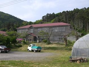 赤い屋根で木造2階建ての校舎、手前の駐車場に停まっている黒い乗用車と白い軽トラックの写真