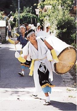 鉢巻、白い和服、前掛けを身につけて草履をはいた男性が大きな太鼓を背負って手にはばちをもって走っている写真