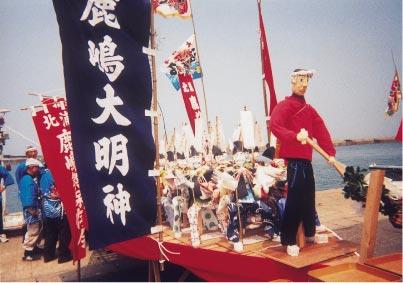 旗が周りにつけられた船のようなものに乗っている、櫂のような棒を持った人形と、そのまわりに立つ法被を着た男性たちの写真