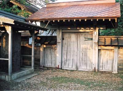 木造の両開きの扉の上に赤いひさしがついた渡部家正門とその左側の屋根のある囲いの中に設置された村法碑の写真