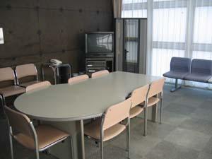 楕円形のテーブルの周りに椅子が置いてあり、壁際にも椅子が設置され、カラオケセットも常備してある練習室(防音室)の写真