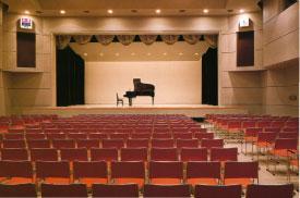 照明のついた舞台中央にピアノと椅子があり、パイプ椅子が並べられている客席後方から写した写真