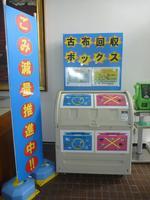 古布回収ボックスの利用について説明のラミネートがボックスの上に貼ってあり、2段のごみ分別があります。ボックスにはマルやバツが貼られその横には「ごみ減量推進中」の文字が書かれたのぼり旗が設置してある写真